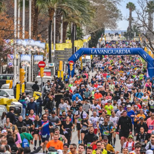 A large crowd of runners starting the Barcelona Marathon under the blue archway, with palm trees lining the street.